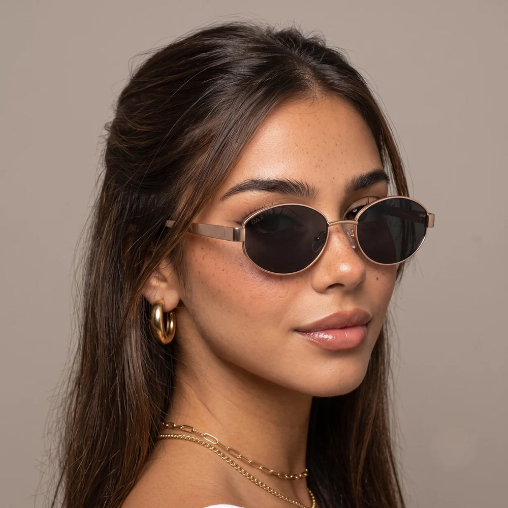 A close-up portrait of a woman wearing oval sunglasses with dark lenses and thin metallic frames, shown against a plain warm gray background.