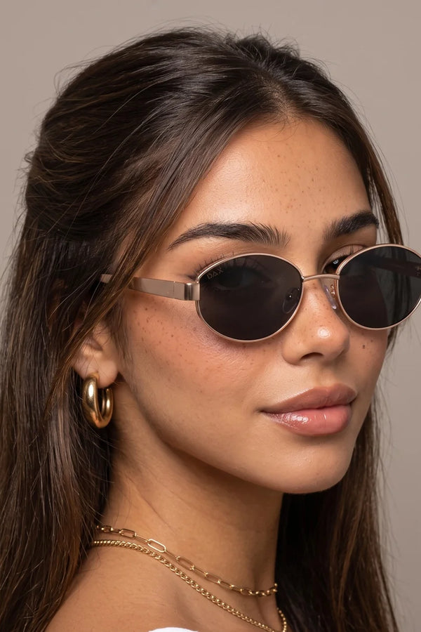 A close-up portrait of a woman wearing oval sunglasses with dark lenses and thin metallic frames, shown against a plain warm gray background.